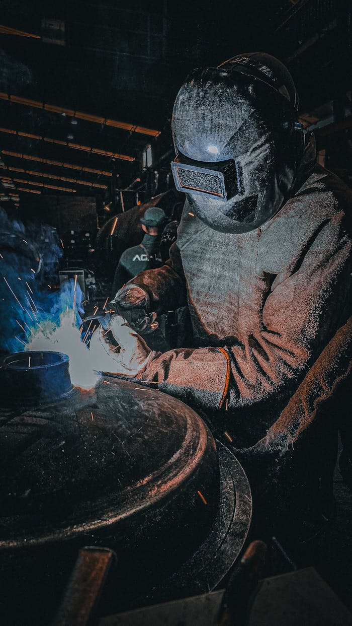 A welder in protective gear working on a metal piece in a workshop with bright sparks.